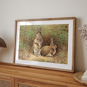 May include: A framed painting of two brown rabbits in a grassy field. The rabbits are standing upright and looking at the viewer. The painting is set against a white background.