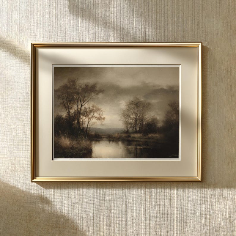 May include: A framed landscape print featuring a sepia-toned scene of trees along a calm river under a cloudy sky. The artwork is encased in a metallic gold frame with a wide, cream-colored mat board, creating a classic presentation for the wall.
