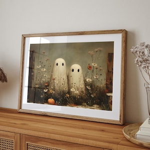 May include: Framed art print featuring two whimsical ghost figures standing amidst wildflowers and a small pumpkin. The artwork has a vintage aesthetic with soft colors and a wooden frame, perfect for Halloween decor.