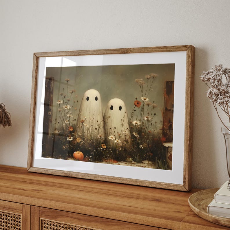 May include: Framed art print featuring two whimsical ghost figures standing amidst wildflowers and a small pumpkin. The artwork has a vintage aesthetic with soft colors and a wooden frame, perfect for Halloween decor.