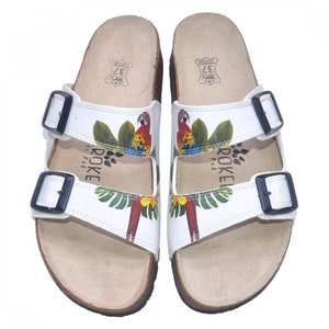 May include: White leather sandals with two adjustable buckles. The sandals feature a tropical print with parrots and palm leaves. The brand name "ROKE" is printed on the straps.