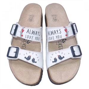 May include: White leather sandals with two adjustable straps and buckles. The sandals have a cork sole and feature a design of birds singing with musical notes and the text "ALWAYS LOVE YOU".