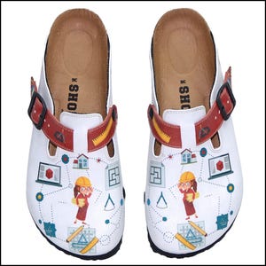Puede incluir: Zuecos de cuero blanco con una correa roja y amarilla y un diseño de herramientas de arquitectura y una mujer con casco y gafas. El texto "SHOES" está impreso en el interior de los zuecos.