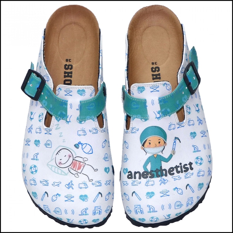 May include: White clogs with a blue and green pattern featuring medical symbols and a cartoon illustration of an anaesthetist. The clogs have a black strap and the text "anesthetist" is printed on the right shoe.