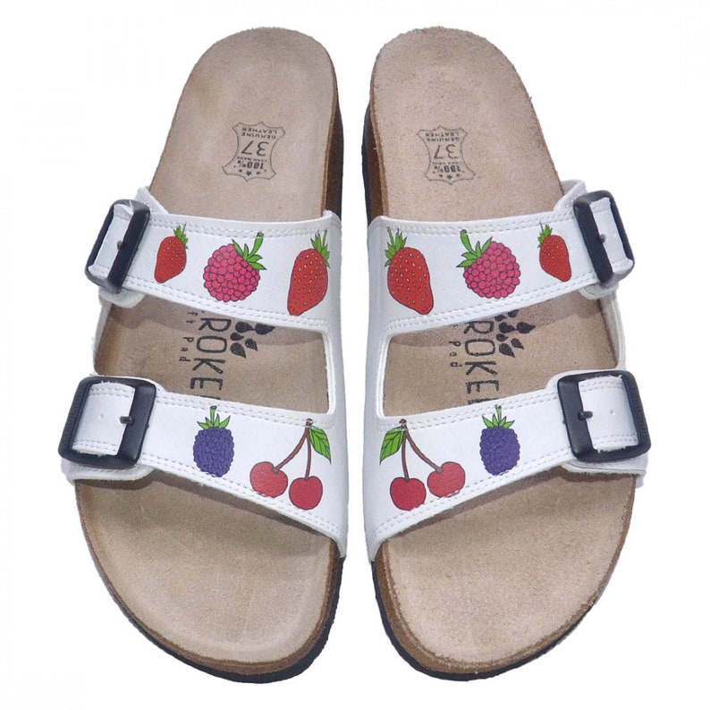 May include: White leather sandals with two adjustable straps and a fruit print. The sandals have a cork footbed and are labeled "ROKE" with the text "100% Leather" on the straps.