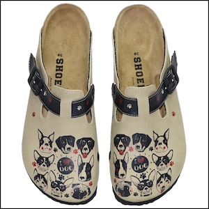 May include: A pair of beige clogs with a black strap and buckle. The clogs are decorated with various dog faces and paw prints. The text "I LOVE DOG" is also featured on the clogs.