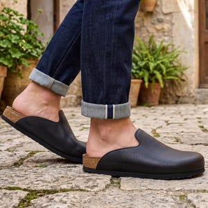 Könnte beinhalten: Ein Paar schwarze Schlupfschuhe aus Leder mit strukturierter Narbung und einer Sohlenkante aus Kork. Das Schuhwerk verfügt über eine abgerundete Zehenpartie und ein offenes Fersendesign, kombiniert mit einer dunkelblauen Jeans mit sichtbarem Umschlag.