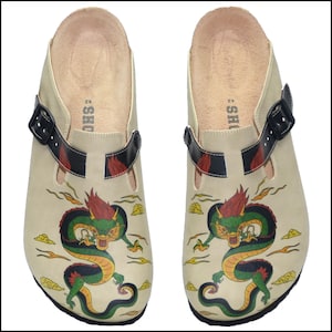 May include: A pair of beige clogs with a black strap and buckle. Each clog features a colorful dragon design with green, red, and yellow accents. The dragon is surrounded by stylized clouds. The clogs have a contoured footbed.