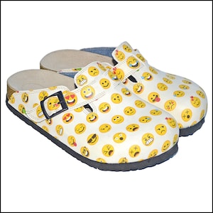May include: A pair of white clogs covered in yellow emoji faces. The clogs have a black sole, a buckle closure, and a strap across the top. The footbed is a light tan color.