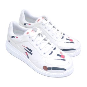 May include: White sneakers with makeup-themed illustrations. The shoes feature drawings of lipstick, mascara, nail polish, and makeup brushes. Small red hearts are also scattered across the white leather surface.