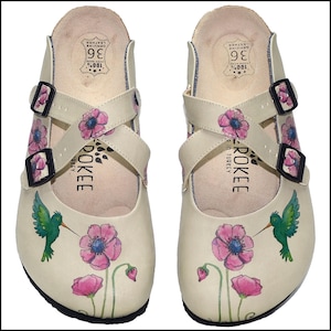 Könnte beinhalten: Paar cremefarbene Lederclogs mit Blumen- und Kolibri-Designs. Die Clogs haben rosa Blumen und grüne Kolibris, verstellbare Riemen und schwarze Schnallen. Größe 36. Der Markenname "ROKEE" ist auf der Seite aufgedruckt. Hergestellt in der Türkei.