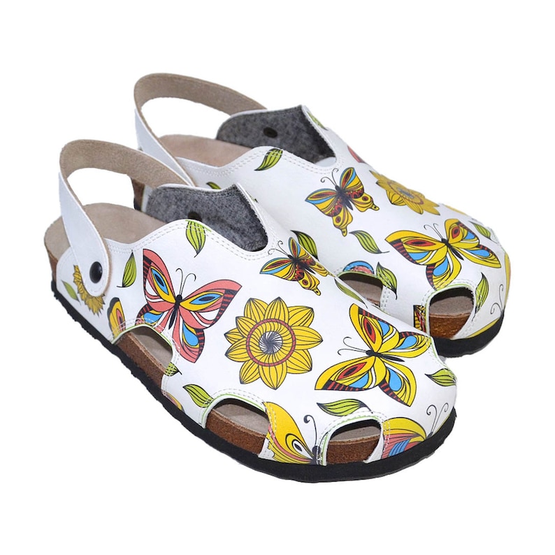 K&ouml;nnte beinhalten: Wei&szlig;e Ledersandalen mit Schmetterlings- und Sonnenblumenmuster. Die Sandalen haben eine Korksohle und einen verstellbaren Riemen.