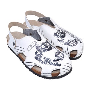Puede incluir: Sandalias de cuero blanco con un diseño de gato de dibujos animados en blanco y negro. El gato dice "¡Boo!" en un globo de pensamiento. Las sandalias tienen una suela de corcho y una correa trasera.