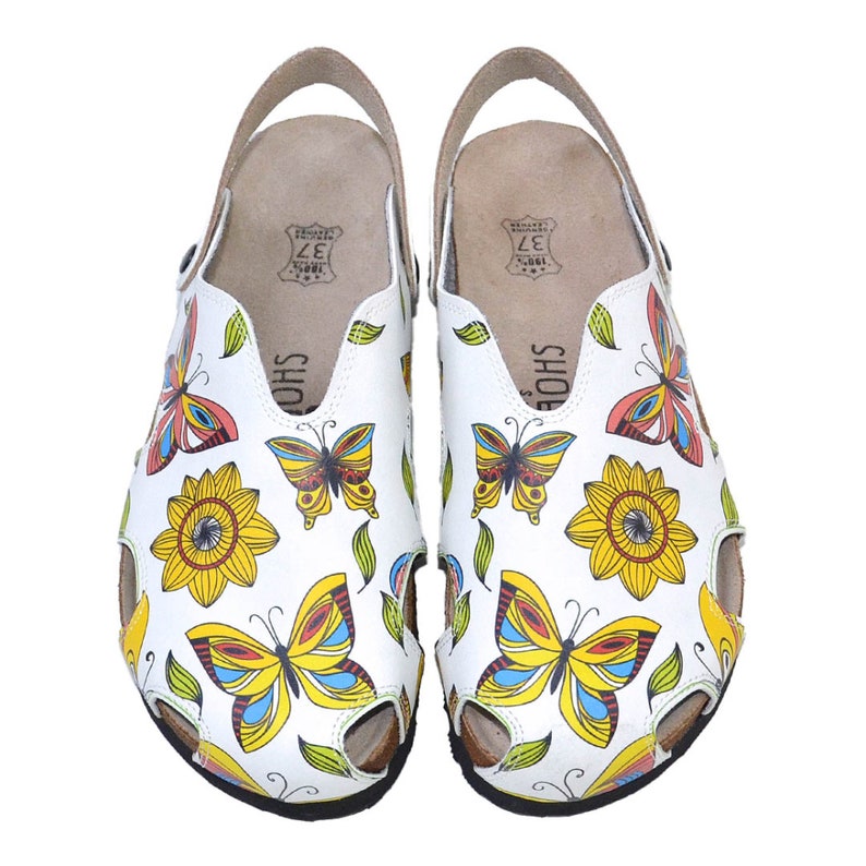 K&ouml;nnte beinhalten: Ein Paar wei&szlig;e Ledersandalen mit einem Schmetterlings- und Sonnenblumenmuster. Die Sandalen haben einen braunen Lederriemen und eine schwarze Sohle.