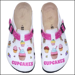 Pode incluir: Tamancos brancos decorados com ilustrações coloridas de cupcakes e a palavra "CUPCAKES" em rosa. Os tamancos têm tiras rosa com fivelas pretas e uma palmilha castanha. Tamanho 37.