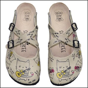 Könnte beinhalten: Beige Lederclogs mit Katzen- und Gitarrenmuster. Die Clogs haben gekreuzte Riemen mit schwarzen Schnallen und den Text "ROKEE TURKEY". Größe 40.