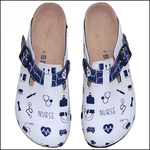 Puede incluir: Un par de zuecos blancos con símbolos médicos azules y la palabra "NURSE" impresa en ellos. Los zuecos tienen una correa negra y una plantilla marrón.