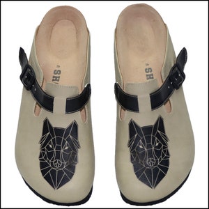 May include: A pair of beige leather clogs with a black geometric wolf design and a black adjustable strap. The clogs have a brown leather insole and the text "SH" is printed on the insole.