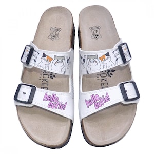 May include: White leather sandals with a cork sole and a double buckle fastening. The sandals feature a cartoon print of cats with the text "hello cutie!" on the front.