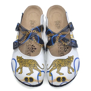May include: White leather clogs with a leopard print design. The clogs feature black straps with gold chain and blue accents. The footbed is brown, and the clogs have a buckle closure. The size is 36.