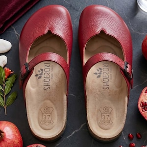 May include: A pair of red leather clogs with a textured finish, featuring a rounded toe, a single adjustable strap with a black buckle across the instep, and a beige contoured footbed stamped with the brand name SHOEROKI and size 37.