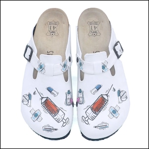 May include: White leather clogs with medical-themed illustrations. Each shoe features a syringe, pills, bandages, and other medical symbols. The clogs have adjustable straps and a cork footbed. The size is marked as 41.