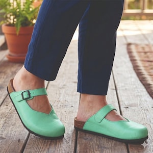 May include: A pair of light mint green leather clogs featuring a rounded toe, a V-shaped cutout at the instep, and an adjustable strap across the top with a dark metal buckle. The footwear has a cork-textured midsole and a dark brown outsole.