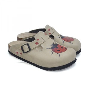 May include: Beige leather clogs with a red ladybug design and black buckle. The clogs have a cork insole and a black rubber outsole.