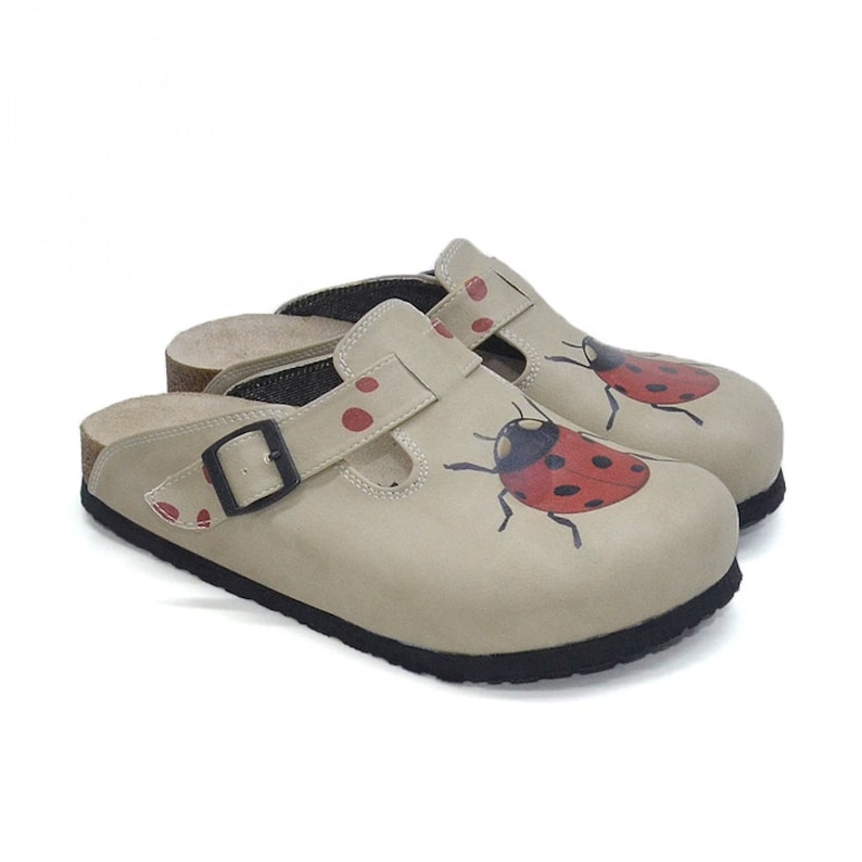 May include: Beige leather clogs with a red ladybug design and black buckle. The clogs have a cork insole and a black rubber outsole.