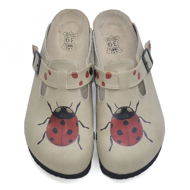 May include: A pair of beige leather clogs with a ladybug design. The clogs have a buckle strap and red polka dots. The size 36 is printed on the inside of the clogs.