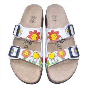 May include: White leather sandals with two adjustable straps and a floral print featuring smiling flowers. The sandals have a cork footbed and are labeled "Jokeee Pad" and "37".