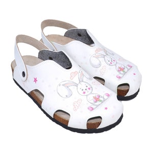May include: White leather clogs with a bunny rabbit design. The clogs have a cork sole and a strap that goes around the ankle.
