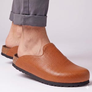May include: A pair of tan brown leather clogs featuring a textured, pebbled surface, a rounded toe, and a cork-style midsole. The footwear has a black, ridged rubber outsole and a slip-on design that leaves the heel exposed for easy wear.