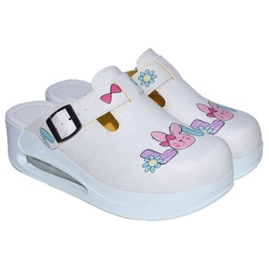 May include: White slip-on clogs with a black buckle and a cartoon bunny design. The clogs have a thick white platform sole.
