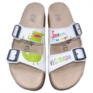 May include: White leather sandals with two adjustable straps and a green dinosaur graphic on one sandal and the text "Sorry, I'm not a vegetarian" on the other sandal.