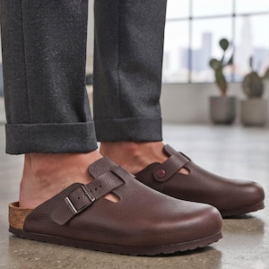 Puede incluir: Un par de zuecos de cuero marrón oscuro con acabado texturizado, punta redondeada, correa ajustable en el empeine con hebilla metálica y entresuela color corcho. El calzado se lleva puesto con pantalones de color gris oscuro.