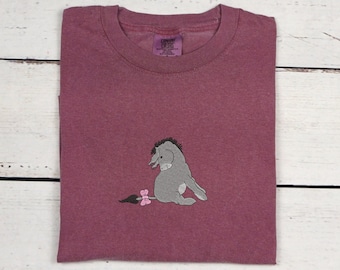 Puede incluir: Una camiseta de color granate con una imagen bordada en gris de Eeyore de Winnie the Pooh. La camiseta tiene cuello redondo y mangas cortas.