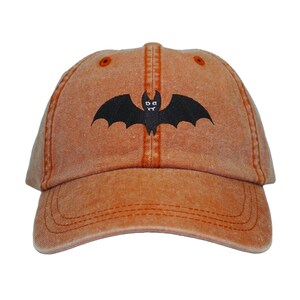May include: A bright orange baseball cap with a black bat graphic. The bat has two white eyes.