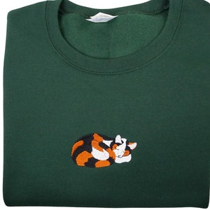 May include: A dark green sweatshirt with an embroidered image of a calico cat curled up sleeping. The cat is orange, black, and white.
