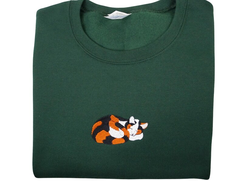 May include: A dark green sweatshirt with an embroidered image of a calico cat curled up sleeping. The cat is orange, black, and white.