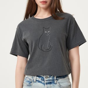 May include: Dark gray t-shirt featuring a minimalist black cat outline. The cat is sitting with its tail curled. The shirt has a classic crew neck and short sleeves. The model is wearing blue jeans.