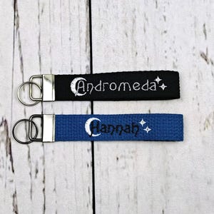 May include: Two fabric keychains with silver-tone metal hardware and split rings. One black strap features the embroidered white text Andromeda with a crescent moon and stars. One blue strap features the embroidered white text Hannah with a crescent moon and stars.