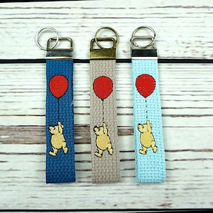 May include: Three keychains with a cartoon bear holding a red balloon. The keychains are blue, gray, and light blue. Each keychain has a silver ring and a rectangular fabric strap.