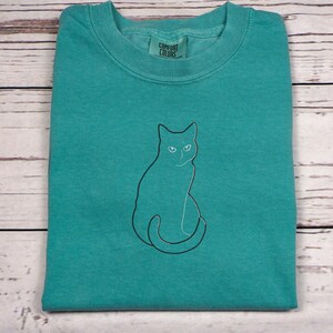 May include: A folded teal-colored t-shirt with a black outline of a cat. The cat is sitting upright with its tail curled. The shirt has a crew neck and a tag that reads "Comfort Colors". The shirt is on a light wood-grain surface.
