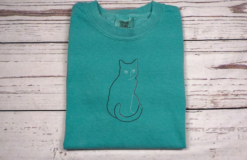 May include: A folded teal-colored t-shirt with a black outline of a cat. The cat is sitting upright with its tail curled. The shirt has a crew neck and a tag that reads "Comfort Colors". The shirt is on a light wood-grain surface.
