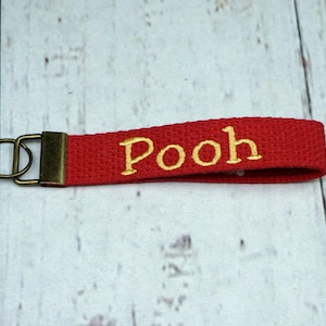 May include: A red fabric keychain with the embroidered word "Pooh" in gold lettering. The keychain has a gold-colored metal clasp and a loop for attaching keys. The background is a light wood grain.