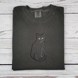 May include: Dark grey t-shirt with a black cat outline embroidered on the front. The cat is sitting with its tail curled. The shirt has a crew neck and short sleeves. The tag reads "Comfort Colors" and "L G G".
