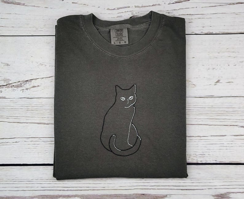 May include: Dark grey t-shirt with a black cat outline embroidered on the front. The cat is sitting with its tail curled. The shirt has a crew neck and short sleeves. The tag reads "Comfort Colors" and "L G G".