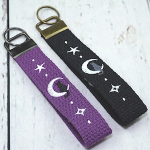 May include: Two keychains with a celestial theme. One is purple, the other black. Both have white embroidered stars, moons, and a cat silhouette. Each keychain has a metal ring and a fabric loop.
