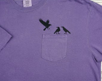 May include: Purple t-shirt with a pocket featuring a black crow design. The design includes three crows, with one in flight. The shirt is made of a soft material and has a classic crew neck.